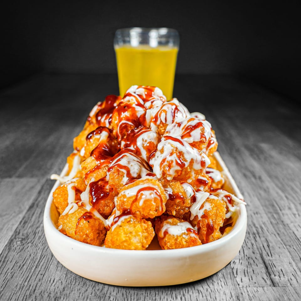 A white bowl of crispy tater tots drizzled with white and BBQ sauces, with a drink in the background.