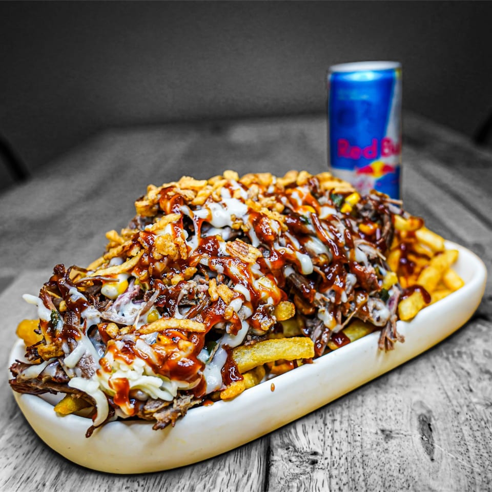 A large platter of KK'S FRIES topped with meat, sauces, and crispy onions, with a Red Bull can.